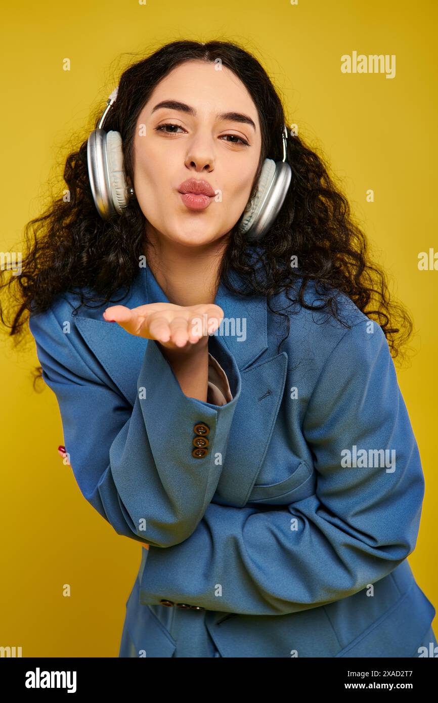 Eine junge, brünette Frau mit lockigem Haar macht einen lustigen Ausdruck, während sie Kopfhörer in einem Studio mit gelbem Hintergrund trägt. Stockfoto