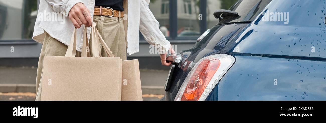 Ein junger, queerer Kerl in stilvoller Kleidung, der Einkaufstaschen neben einem blauen Auto hält Stockfoto