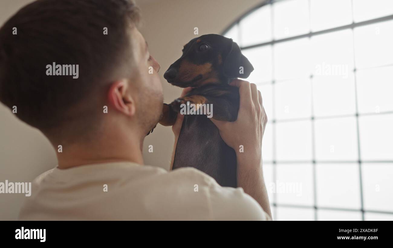 Junger hispanischer Mann, der einen Dackelhund in einem gemütlichen Wohnzimmer hält und einen zärtlichen Moment zwischen dem Haustier und seinem Besitzer zeigt. Stockfoto