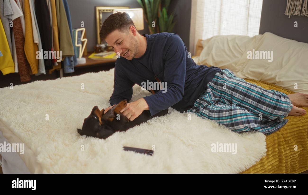 Ein junger hispanischer Mann im Pyjama liegt auf einem Bett in einem gemütlichen Schlafzimmer und spielt mit seinem Haustier-Dackelhund. Stockfoto