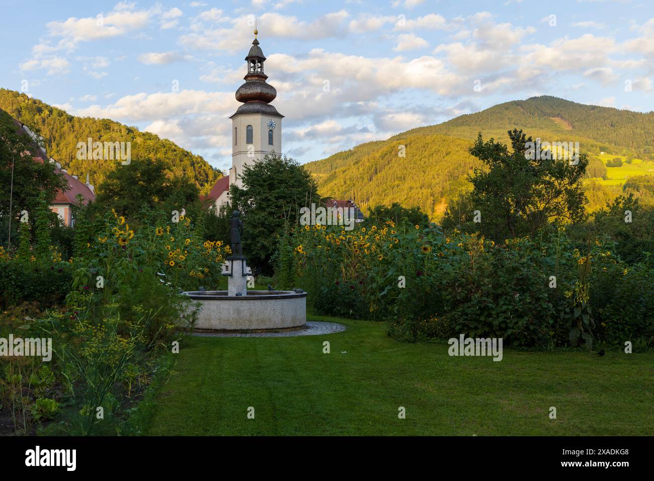 Foto von einem mittelalterlichen Glockenturm und roten Dachhäusern umgeben von grünem Garten mit alpinen Bergen auf einem Hintergrund, alpine Dorflandschaft in einer Sonne Stockfoto