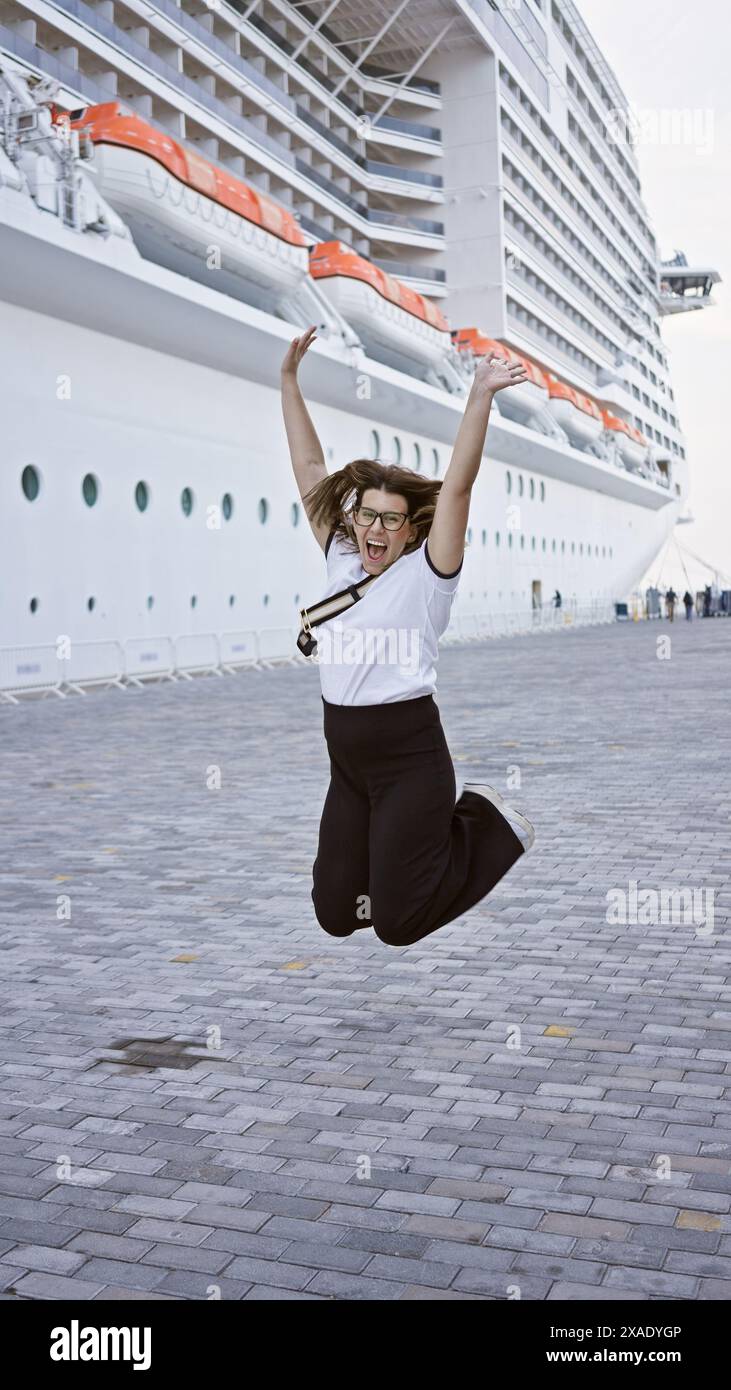 Eine freudige Frau springt vor Begeisterung, bevor sie an Bord eines Kreuzfahrtschiffs für eine Luxusreise geht. Stockfoto