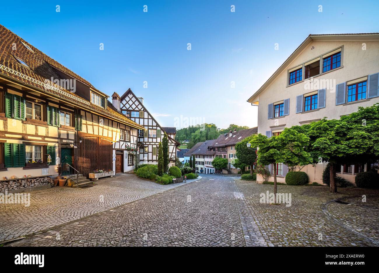 Schweizer mittelalterliches Dorf Regensberg, Platz mit traditioneller Architektur, Kirche, Steinturm, Kopfsteinpflasterstraßen und Grün unter einem klaren Blau Stockfoto