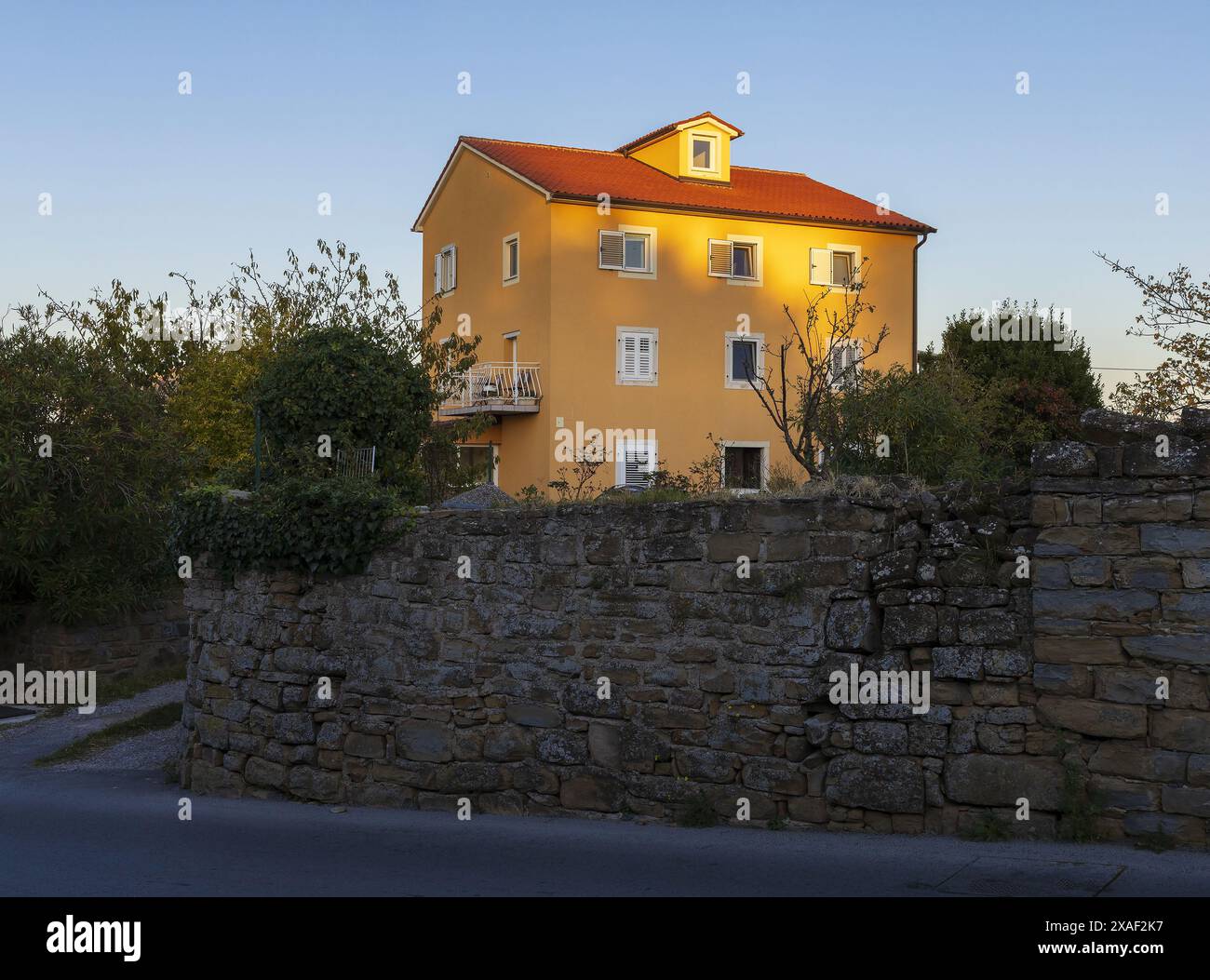 Foto eines gelben Hauses mit weißem Balkon, Fenstern und rotem Dach, umgeben von Garten und Mauer bei Sonnenuntergang im Sommer mit warmem Licht Stockfoto