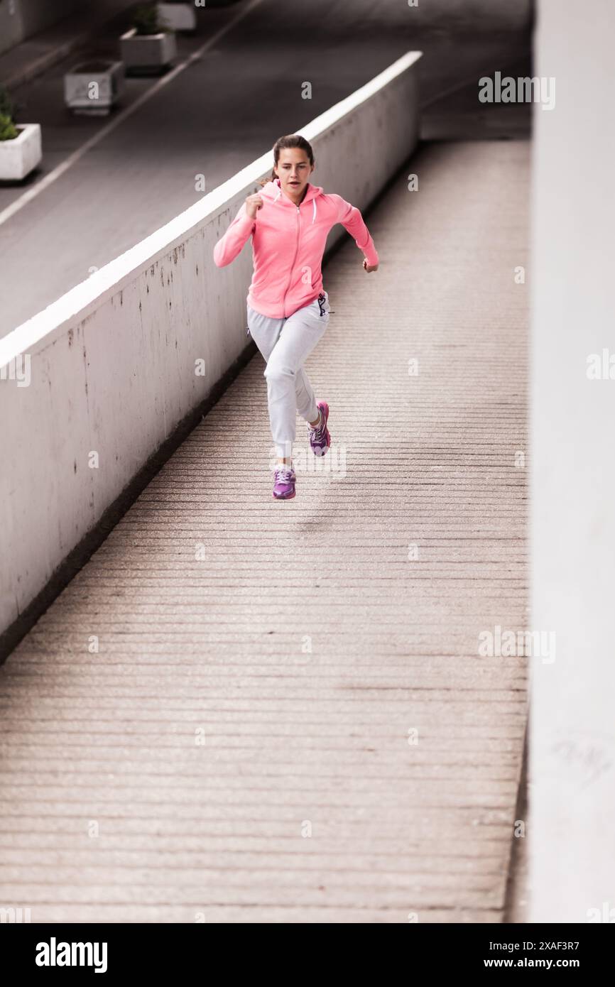 Eine fokussierte junge Läuferin in einer leuchtend pinkfarbenen Jacke sprintet auf einem Betonweg in einer urbanen Umgebung. Perfekte Erfassung von Fitness und Determination Stockfoto