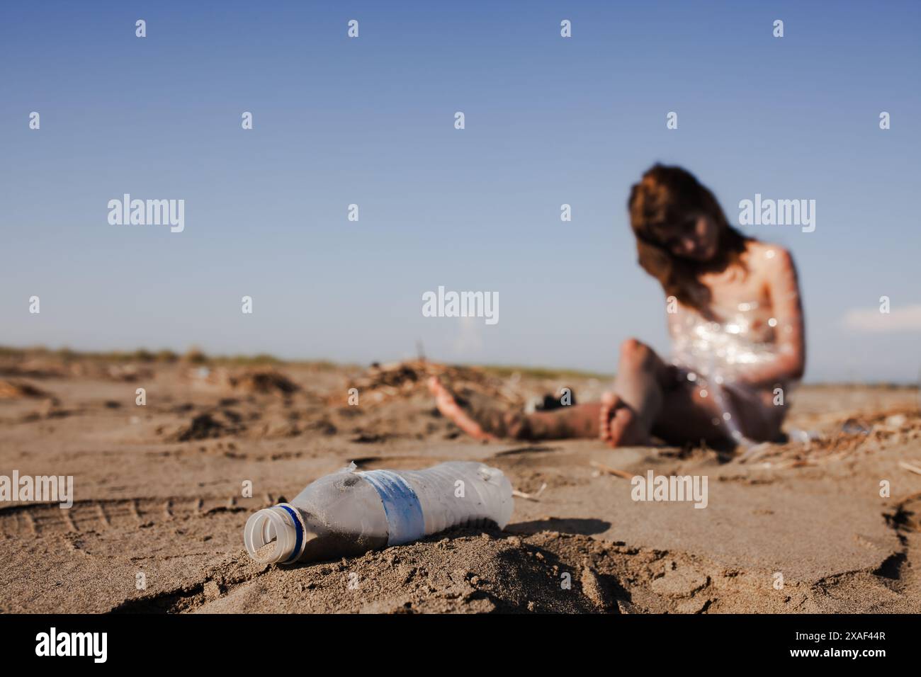 Eine nachdenkliche junge Frau in einem Paillettenkleid denkt über Umweltprobleme nach, während sie neben einer weggeworfenen Plastikflasche an einem einsamen Sandstrand sitzt Stockfoto