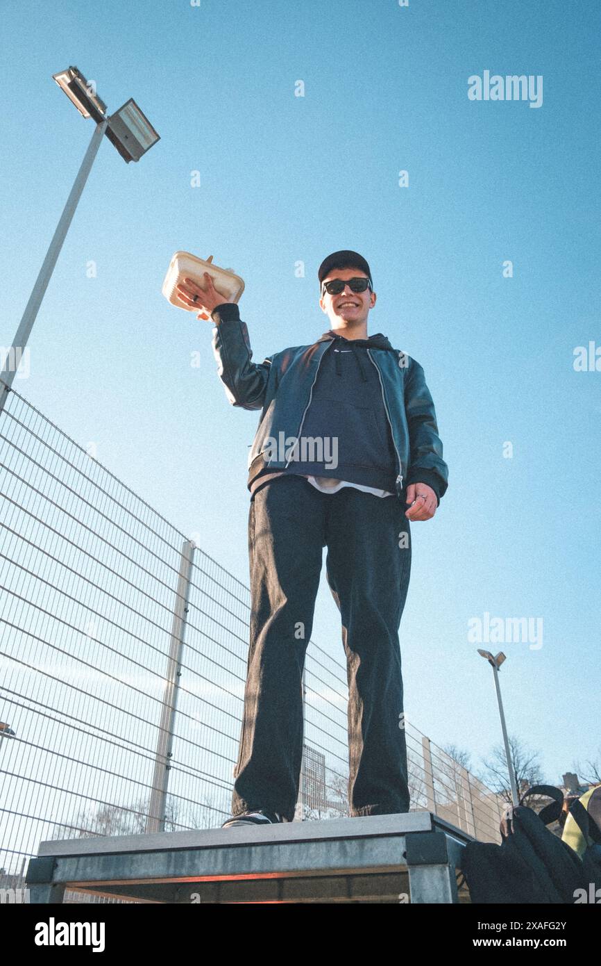 Eine Gruppe von Teenagern sitzt draußen auf einem Basketballfeld mit Ständen, isst Kebab, spielt Basketball, hat Spaß zusammen, hört Musik. Stockfoto