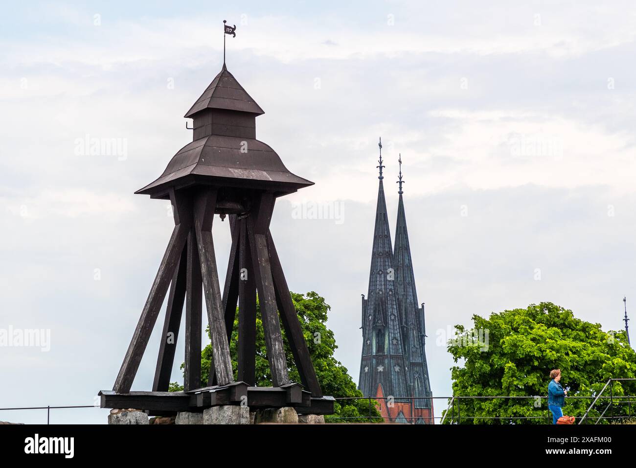 Der hölzerne Glockenturm von 1756 steht auf den Überresten einer Mauer aus dem Schloss, das 1702 niederbrannte, mit den Türmen der Uppsala-Kathedrale, die dahinter aufragen. Blick vom höheren Schlossplatz, ein Passant geht rechts vorbei. Drottning Christinas väg, Uppsala, Uppsala län, Schweden. Stockfoto