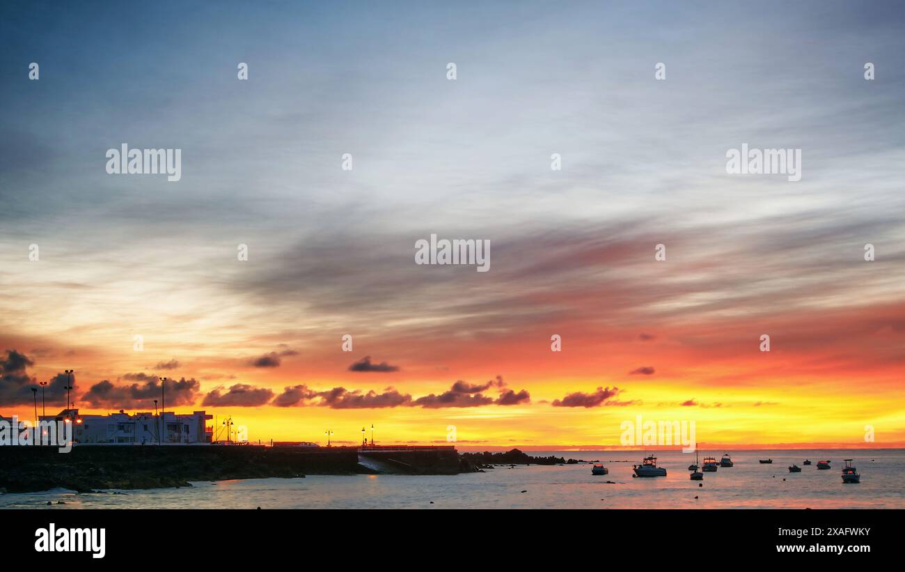 Ein lebendiger Sonnenaufgang über einem Fischerdorf auf den Kanarischen Inseln, mit farbenfrohen Himmeln, verstreuten Wolken und Booten, die im ruhigen Meer verankert sind. Stockfoto