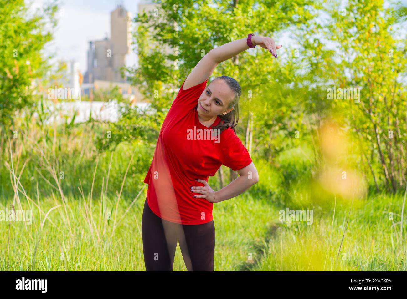 Eine Frau mit rotem Hemd, die sich auf einem grasbewachsenen Feld ausdehnt. Sie hat ihren rechten Arm über dem Kopf und ihre linke Hand auf der Hüfte Stockfoto