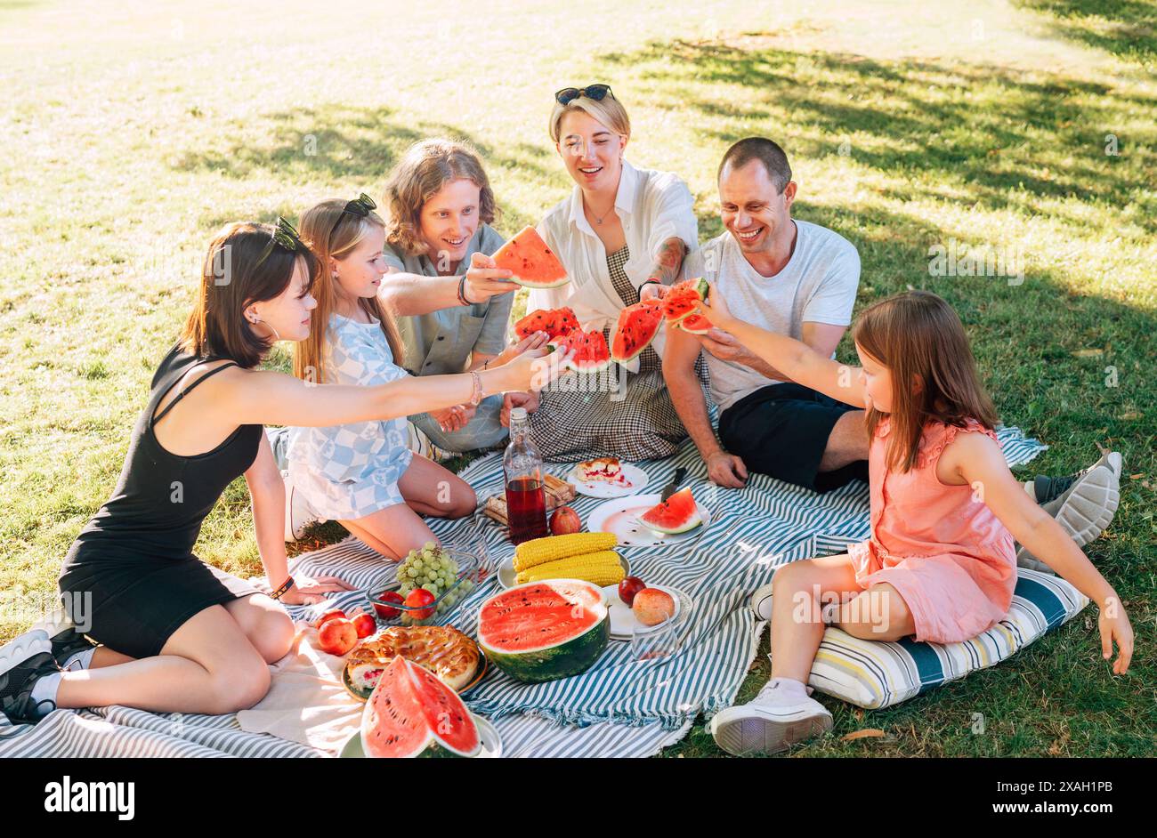 Große kaukasische Familie auf Picknickdecke auf dem grünen Gras im Stadtpark. Sie haben rote, saftige Wassermelonenstücke aufgehauen und sie wie Gläser angeschliffen. Familie Stockfoto
