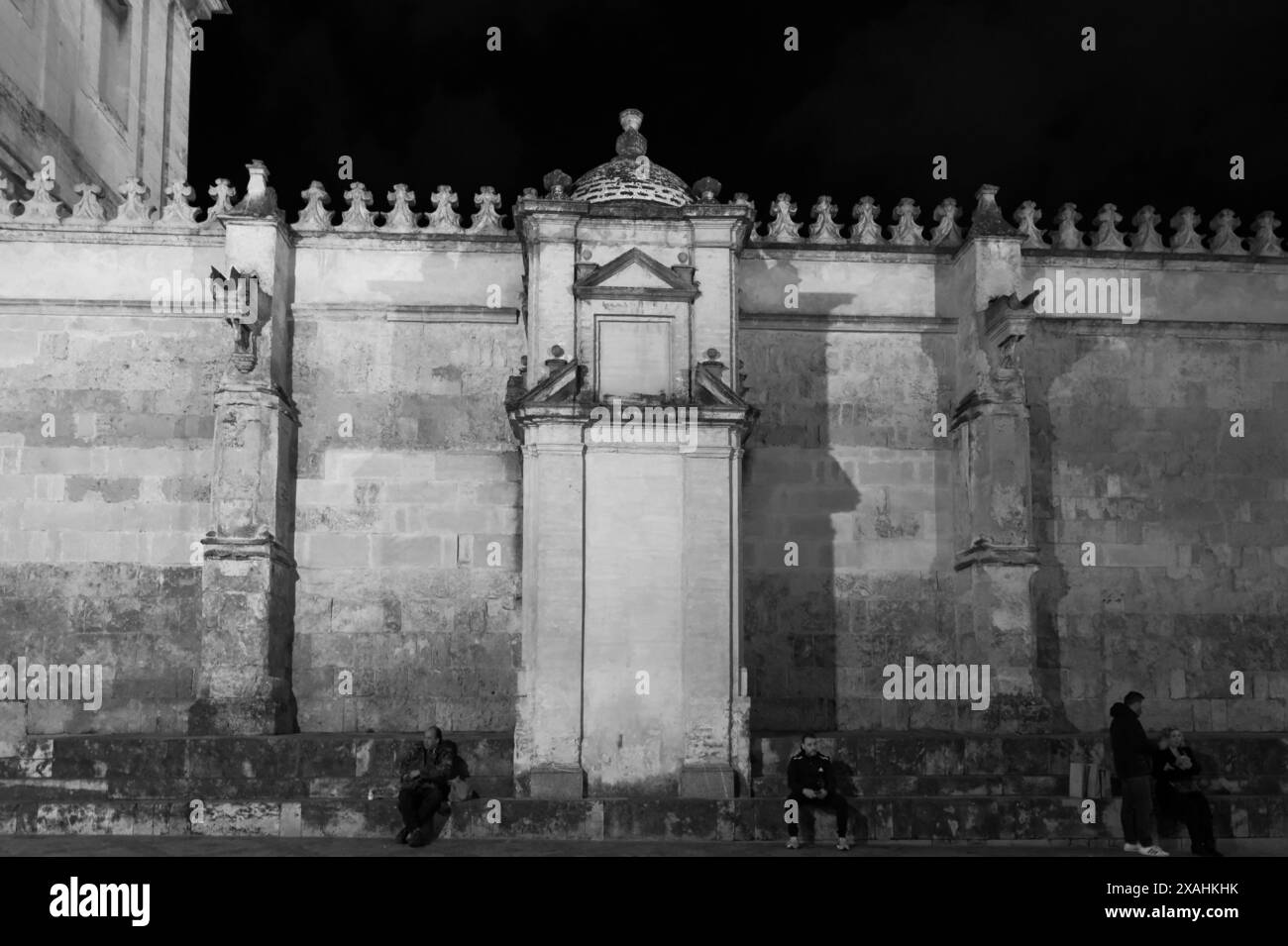 Die historische Steinmauer rund um die Moschee-Kathedrale von Córdoba, die bei Nacht beleuchtet wird, zeigt architektonische Details, während die Menschen sitzen und sich unterhalten Stockfoto