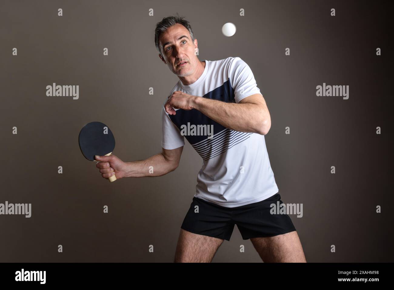 Porträt des konzentrierten Tischtennisspielers, der einen orangen Ball auf grauem, isoliertem Hintergrund trifft. Stockfoto