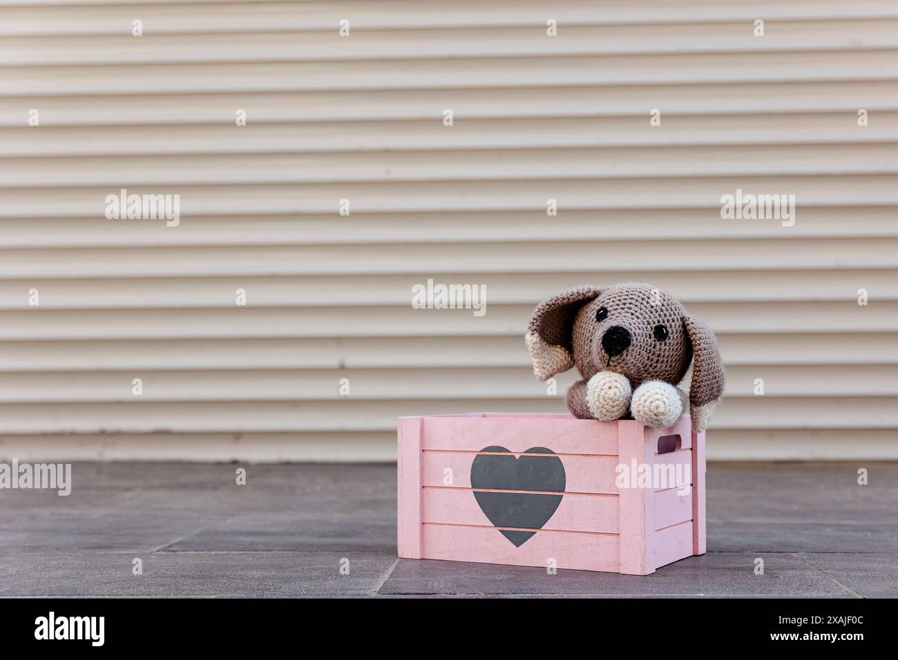 Gestrickter Welpe in rosafarbener Holzkiste mit Herz drauf. Niedlicher, häkelter Spielzeughund mit dem Kopf auf den Pfoten, der vor Trauer aussieht. Übernehmen Sie das Konzept „nicht einkaufen“ Stockfoto