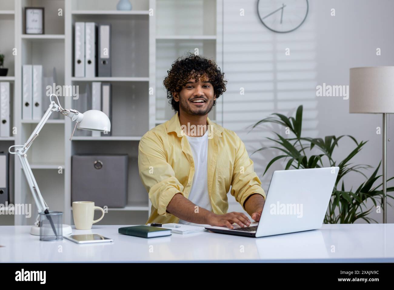 Ein lächelnder junger Mann an seinem Schreibtisch mit einem Laptop in einer hellen und modernen Büroumgebung. Konzept von Arbeit, Glück und Produktivität. Stockfoto
