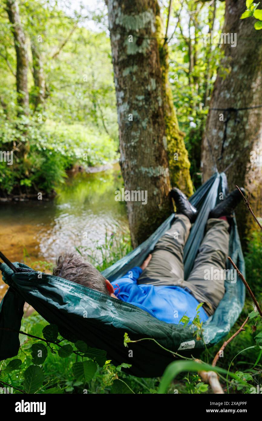 Ein Mann mittleren Alters liegt entspannt in einer Hängematte neben einem kleinen Bach in Devon, England auf einem wilden Campingplatz. Stockfoto