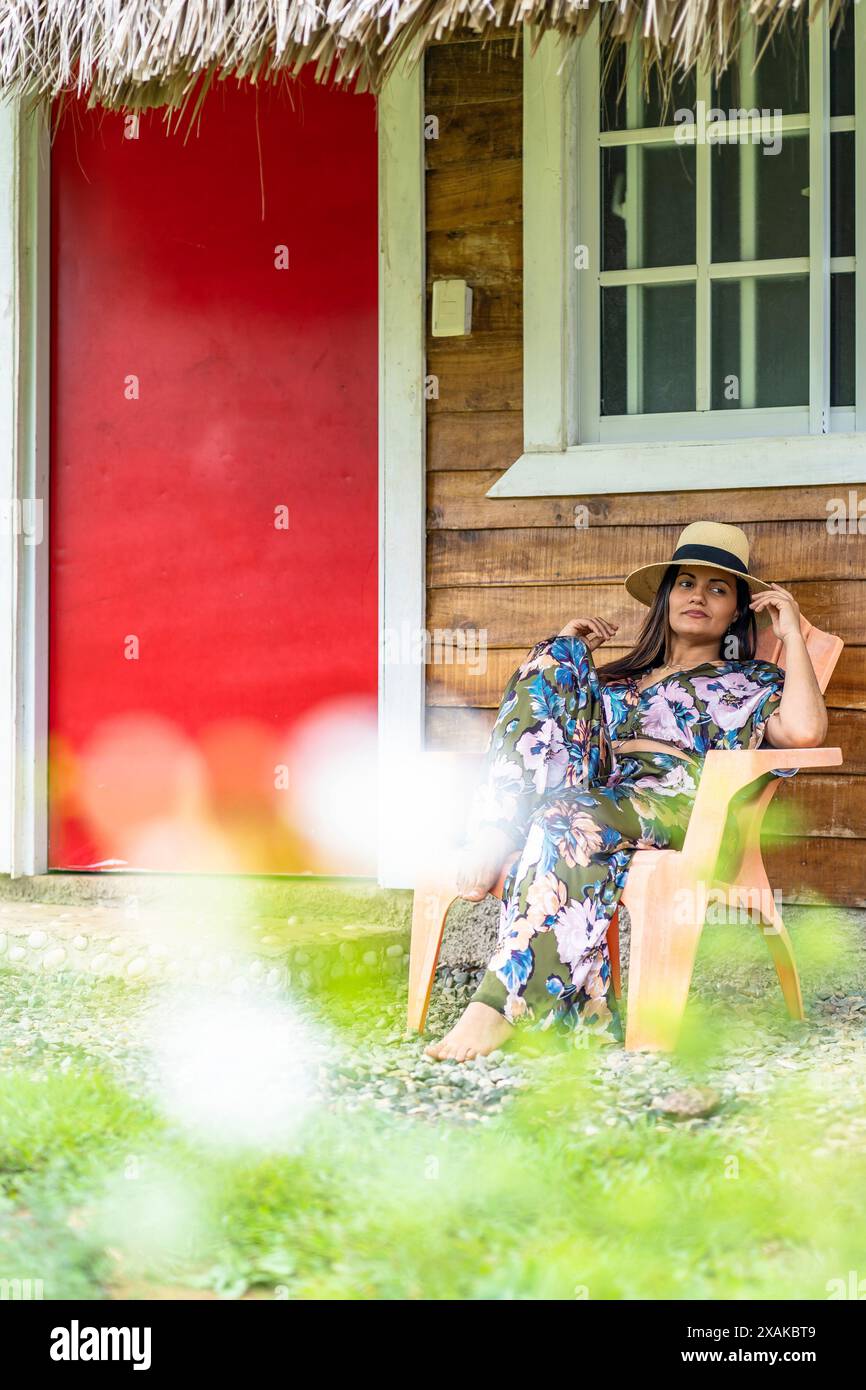 Nordamerika, Karibik, Hispaniola Island, Dominikanische Republik, Monte Plata, Bayaguana, Campo Aventura, hübsche Latina, die auf einem Stuhl vor einem rustikalen Holzhaus sitzt Stockfoto