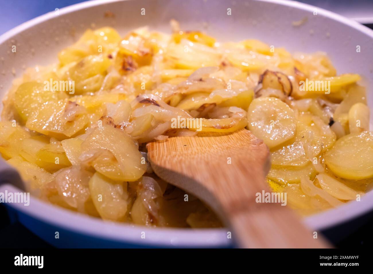 Ein Teller mit frischen Kartoffeln und Zwiebeln wird in einer Pfanne aus weißem Granit gebraten. Holzlöffel für ein biologisches, ökologisches, natürliches, gesundes und köstliches Gericht. I Stockfoto