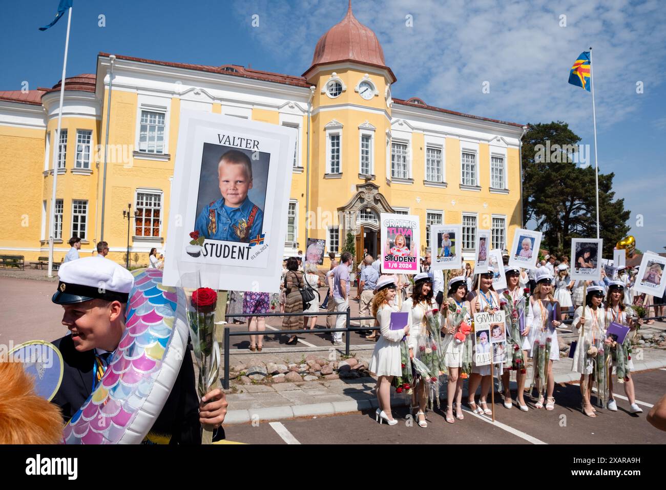 JUNGE MIT BALLON, MÄDCHEN POSIEREN FÜR GRUPPENBILD, Åland ABSCHLUSS, MARIEHAMN: Ein Junge mit Ballon und eine große Gruppe von Mädchen Absolventen posieren für ein festliches Bild mit Blumen und ihren Babyplakaten vor dem Hauptschulgebäude am Åland Graduierungstag 2024 im Åland Lyceum (Ålands Gymnasium) in Mariehamn, Åland Archipel, Ostsee, Finnland – an diesem Tag verlassen die Schüler zum letzten Mal die Schule. Im Rahmen dieser nordischen Tradition werden Plakate mit Babybildern der Absolventen aufgehalten und die Studenten tragen ihre Studentmössa im Matrosenstil. Stockfoto