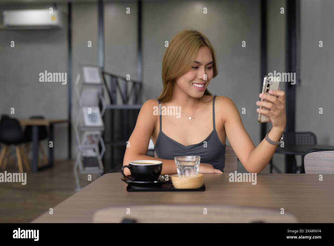 Porträt einer schönen jungen asiatischen Frau, die in einem Café sitzt und in professioneller Kleidung sitzt, während sie ihr Handy benutzt und lächelt Stockfoto