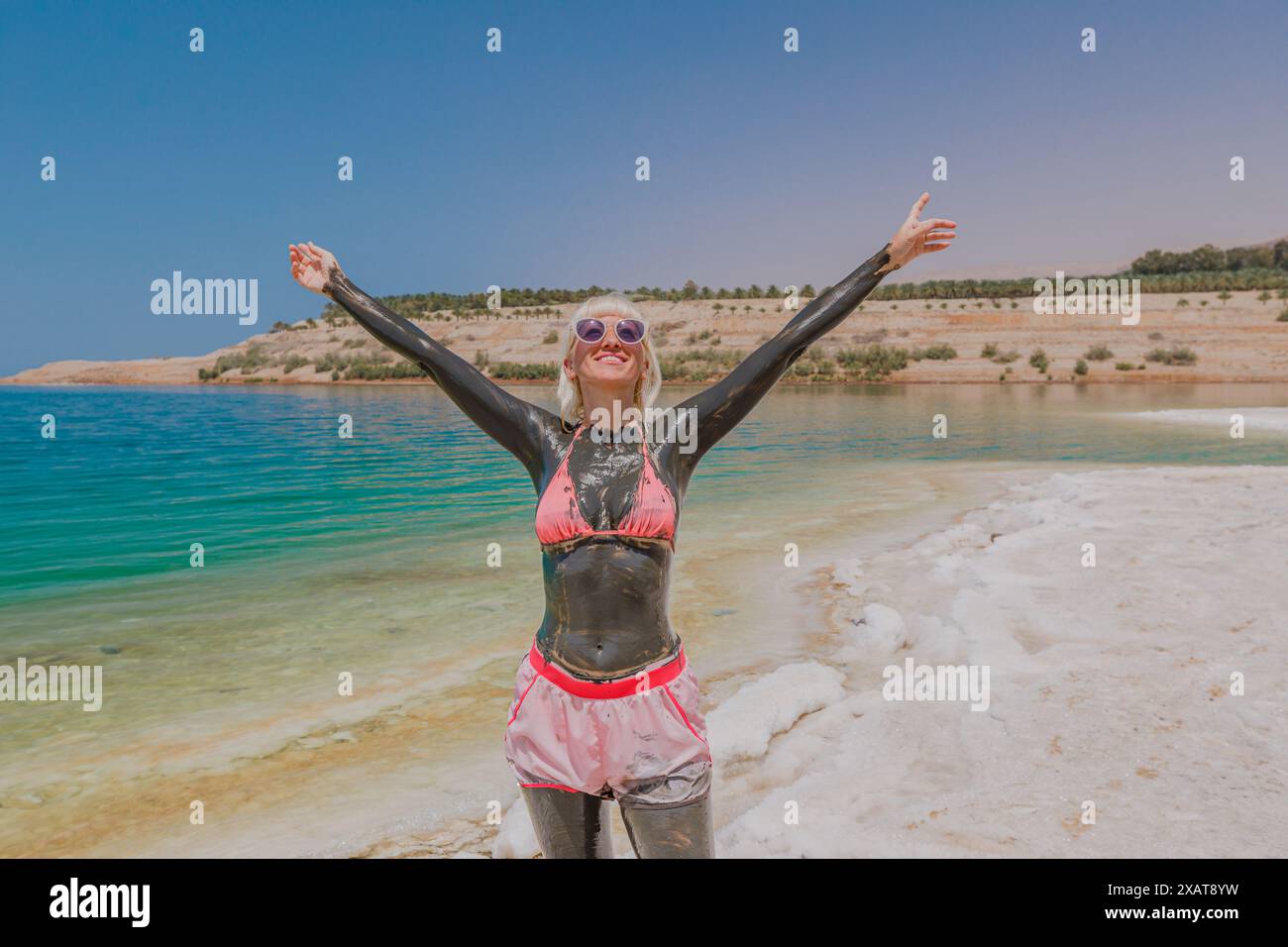 Fröhliche blonde Frau in Sonnenbrille bedeckt mit Schlamm vom Toten Meer, die Thalassotherapie macht, an einer felsigen Küste unter einem klaren blauen Himmel in Jordanien Stockfoto