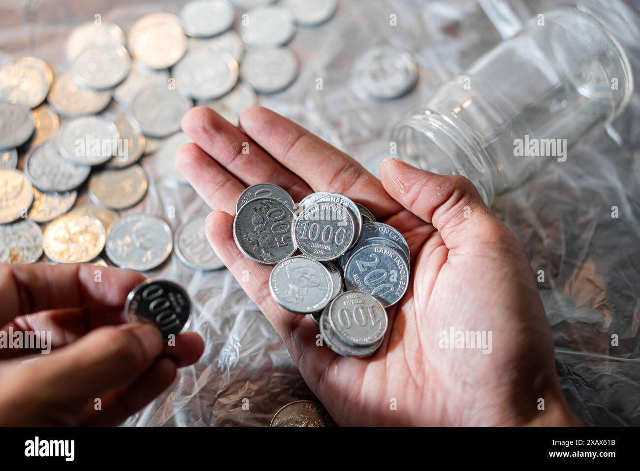 Eine Hand, die mehrere südkoreanische Won-Münzen mit weiteren Münzen und einem leeren Glasgefäß im Hintergrund hält. Stockfoto