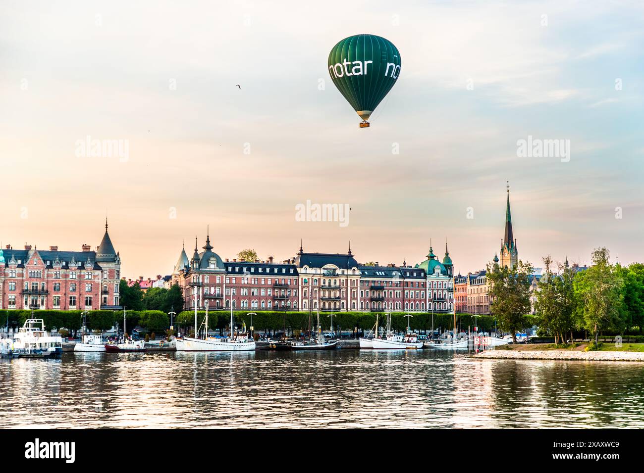 Ein mit Notar gebrandetes Heißluftballon schwebt über den Wasserfrontgebäuden an der Strandvägskajen in Stockholm. Verschlungene Stadthäuser aus dem späten 19. Jahrhundert säumen das Ufer, mit Booten und Segelschiffen im Vordergrund, die vor Anker liegen. Stockholm wird auch als die „Venedig des Nordens“ bezeichnet, aufgrund seiner vielen Wasserwege und Brücken. Strandvägskajen, Stockholm, Stockholms län, Schweden. Stockfoto
