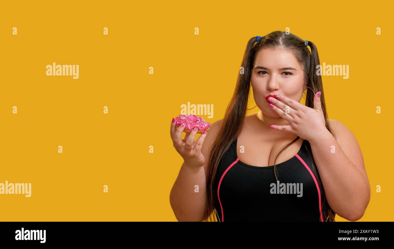 Süßes Leben. Übergewichtige Frau. Rosa glasierter Donut. Niedliches molliges Modell genießt leckeren Donut, der Mund mit Fingern im Studio isoliert auf gelbem Leerraumhintergrund berührt. Stockfoto