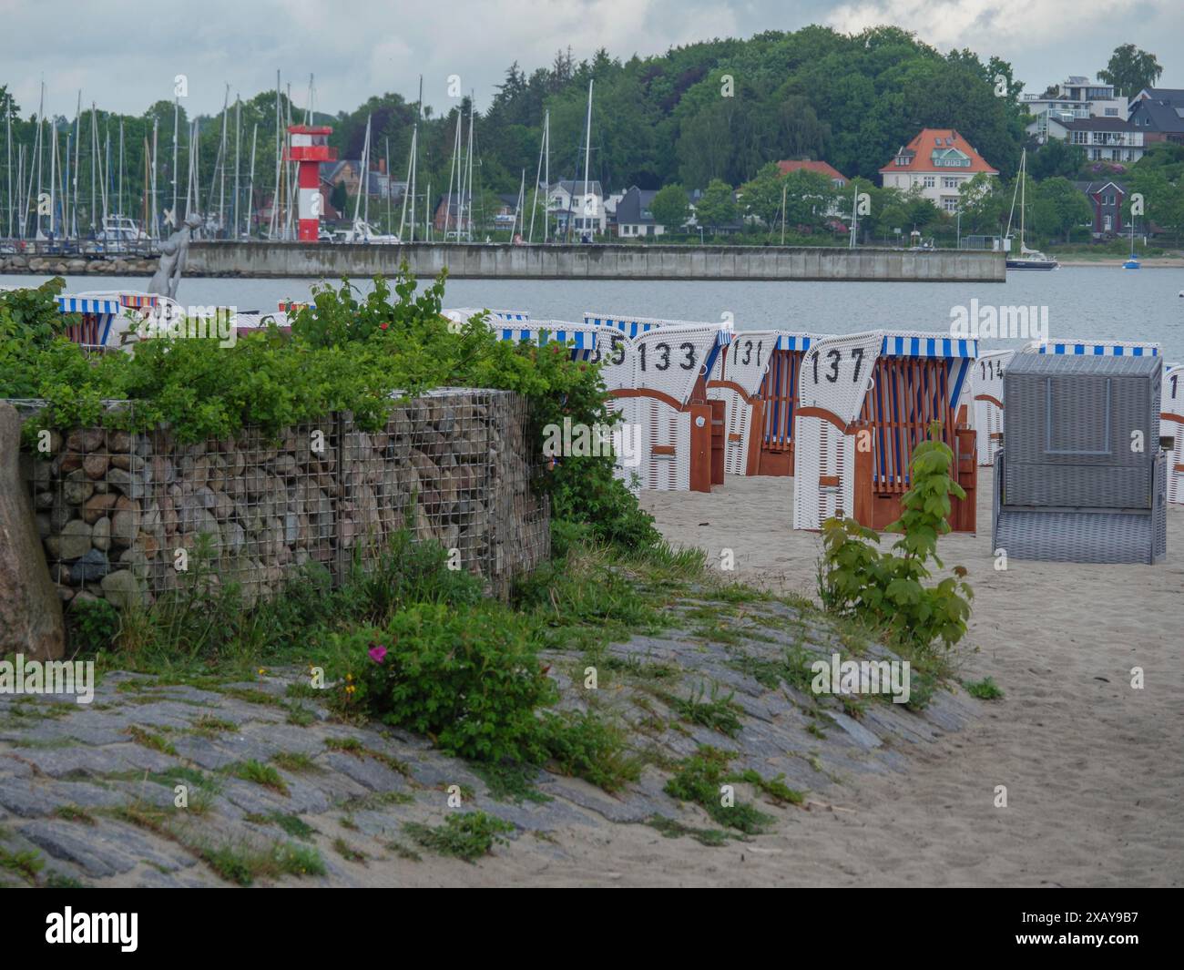 Liegestühle entlang der Küste, mit Blick auf einen Jachthafen und Strandhäuser nebeneinander unter bewölktem Himmel, Eckernförde, Schleswig-Holstein Stockfoto