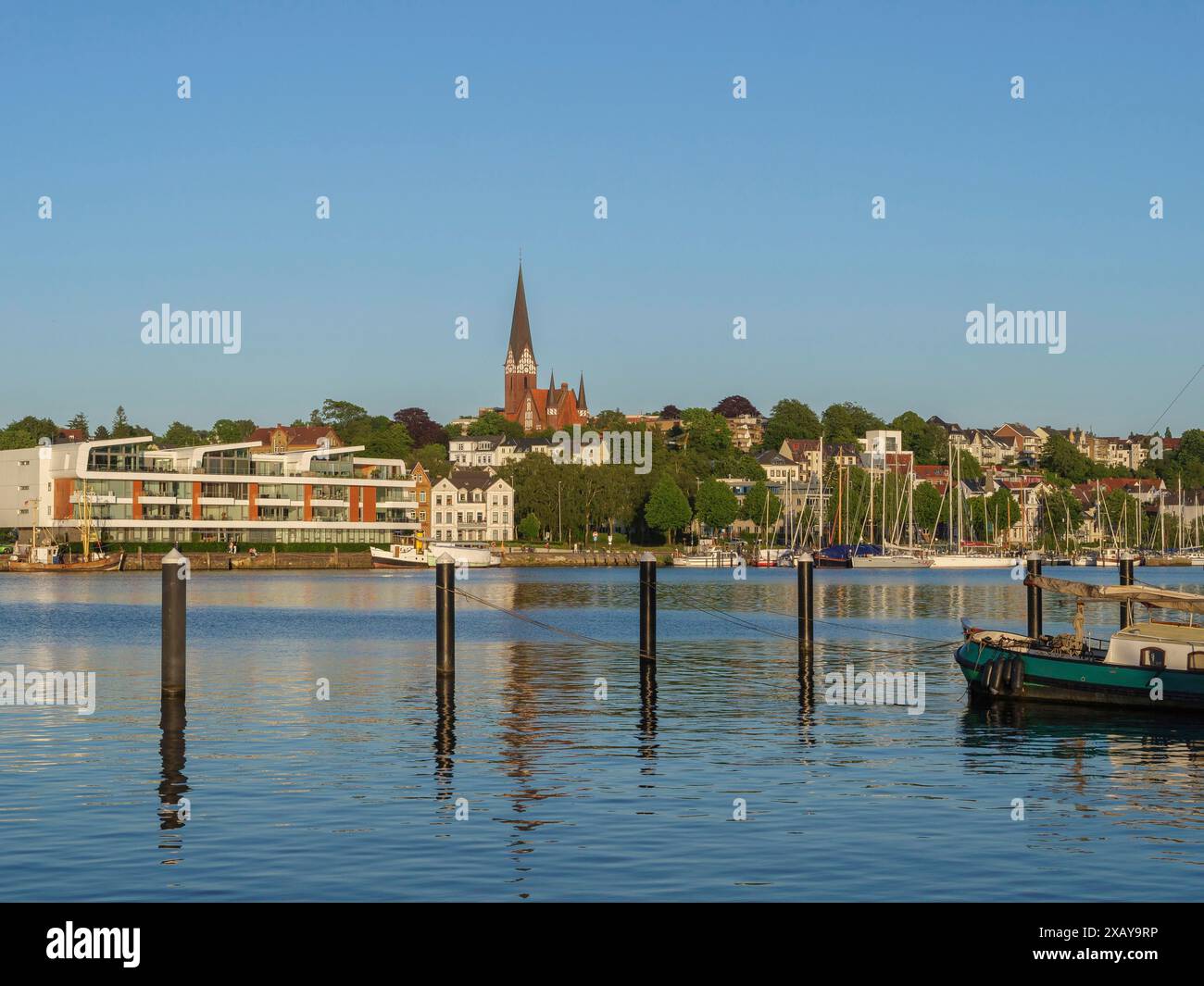 Blick auf die Stadt am Wasser mit Kirche, modernen Gebäuden und Booten vor klaren Pfählen im Morgenlicht, Flensburg, Schleswig-Holstein Stockfoto