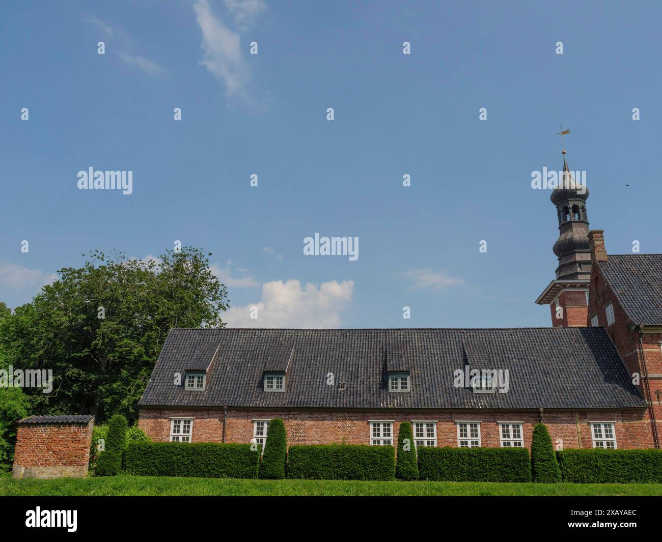 Ein langes Gebäude mit hohem Turm, dunkelgrauem Dach und ordentlich getrimmten Hecken unter hellblauem Himmel, husum, schleswig-holstein, deutschland Stockfoto