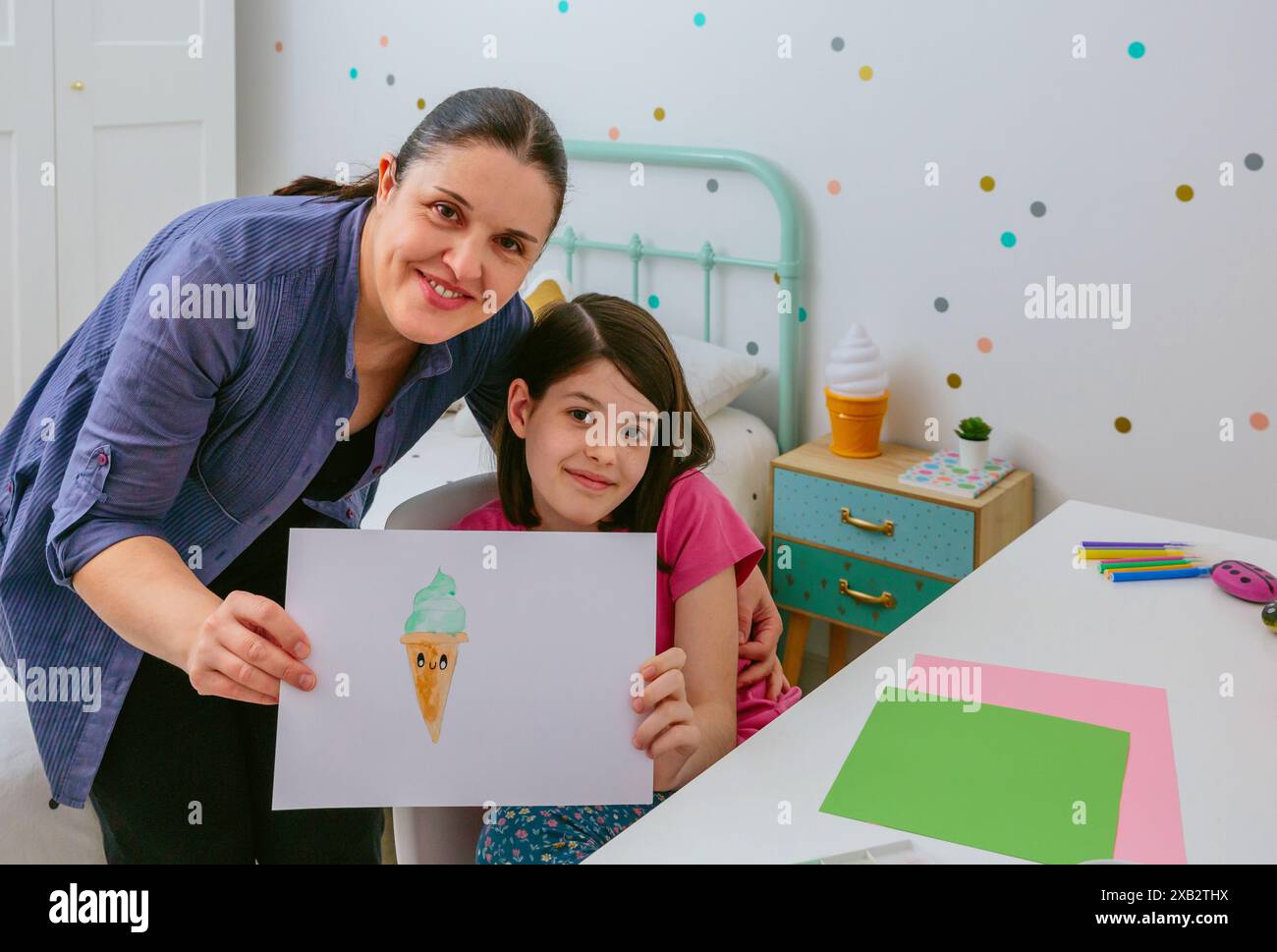 Mutter und Tochter lächeln zusammen, während sie eine Zeichnung einer Eiskugel in einem Schlafzimmer mit Polka-Punktmuster halten Stockfoto