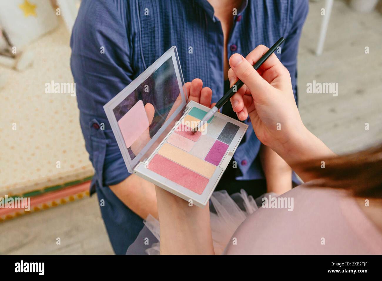 Erwachsene Hände halten eine Schminkpalette, während ein Kind in einem gemütlichen Schlafzimmer eine Farbe mit einem Pinsel wählt Stockfoto