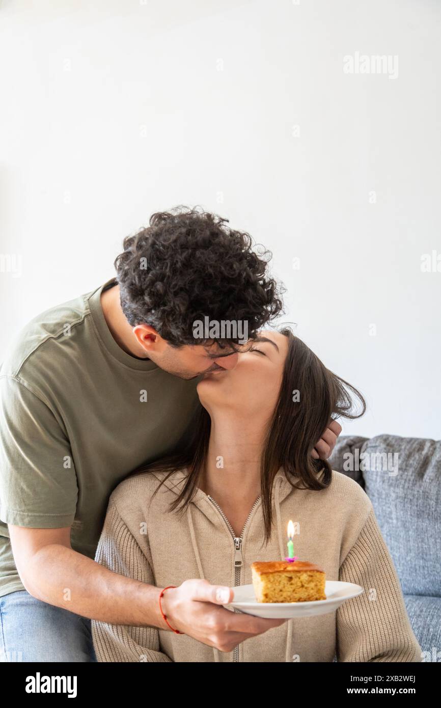 Ein romantischer Moment zwischen einem Mann mit lockigen Haaren und einer Frau, während er sie auf die Lippen küsst und ihr eine Geburtstagskuche mit einer einzelnen Dose überreicht Stockfoto