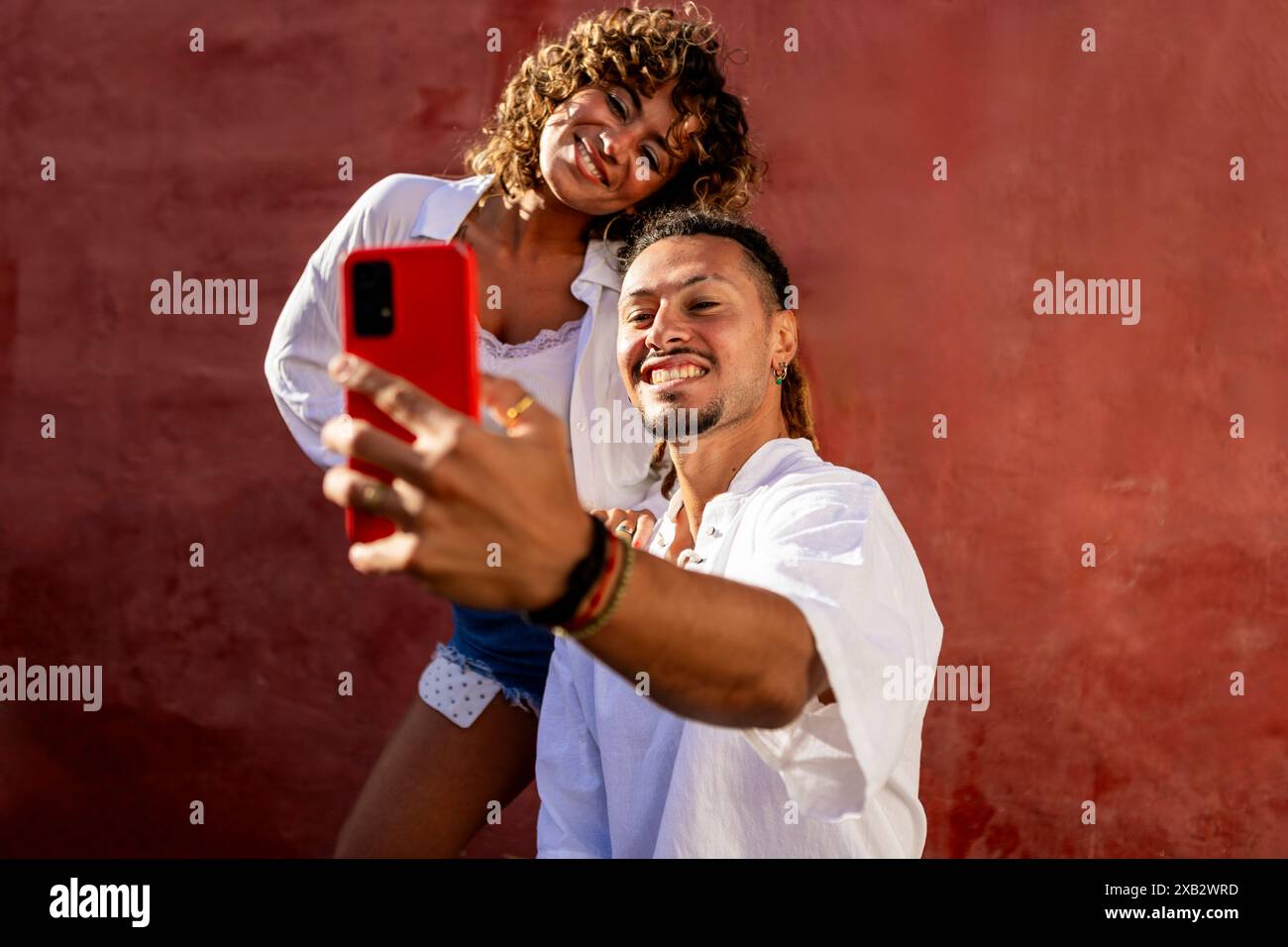 Ein fröhliches junges Paar macht ein Selfie gegen eine leuchtend rote Wand. Mann und Frau, beide in lässigen weißen Outfits gekleidet, teilen einen verspielten Moment Stockfoto