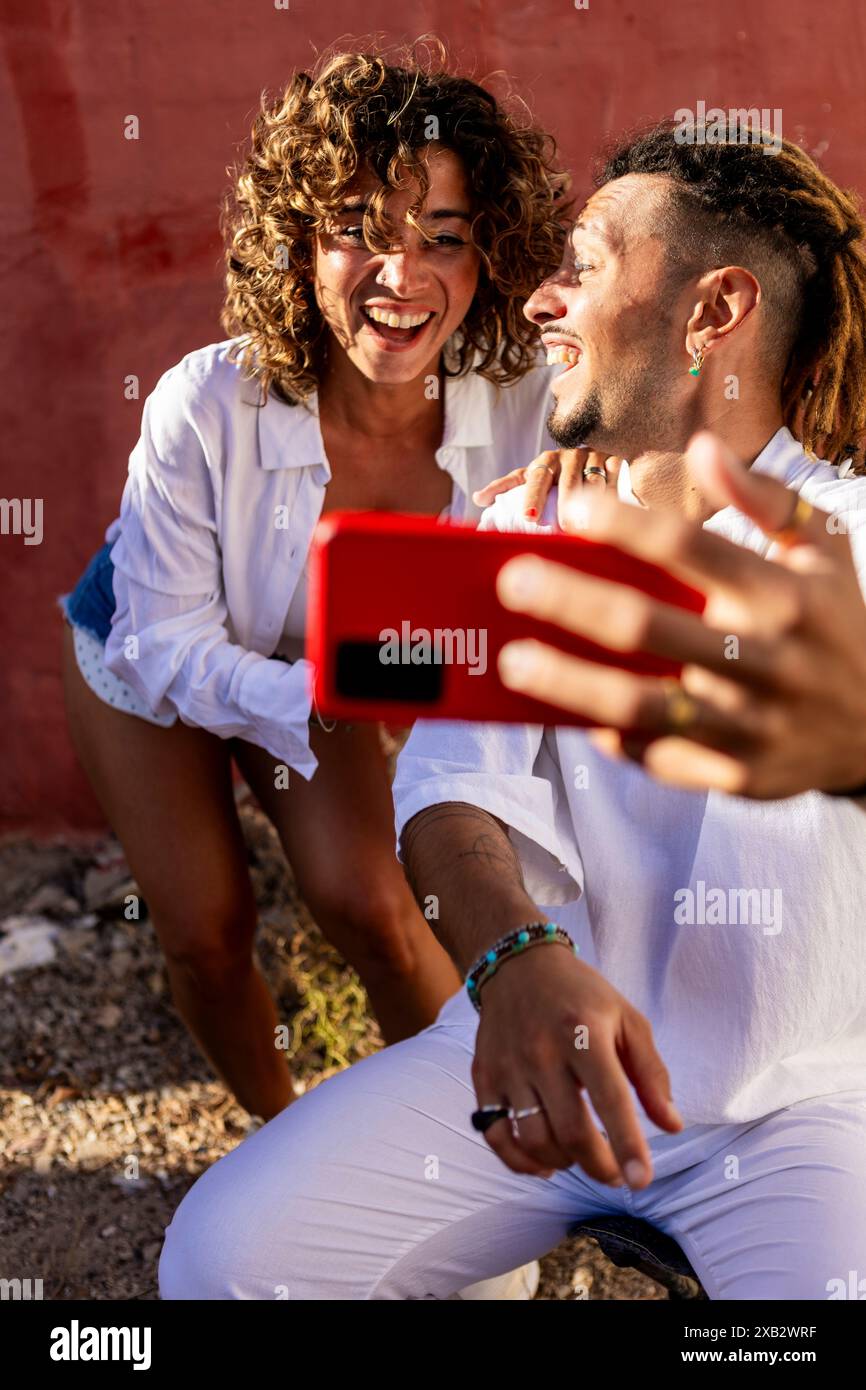 Ein fröhliches junges Paar in lässiger weißer Kleidung genießt einen verspielten Moment, während es draußen ein Selfie gegen eine leuchtend rote Wand macht. Die Frau lacht hell Stockfoto
