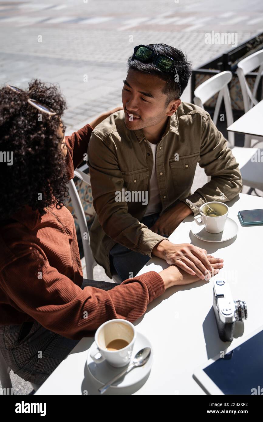 Ein chinesischer Mann und eine hispanische Frau verbringen einen intimen Moment bei einem Kaffee in einem Café im Freien in Madrid, Spanien. Sie halten die Hände und unterhalten sich, fangen sie ein Stockfoto