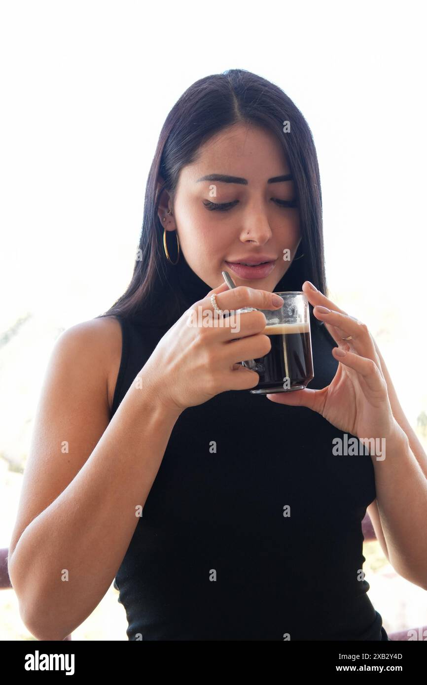 Eine junge Frau in einem schwarzen ärmellosen Oberteil, die den Geschmack von frischem schwarzem Kaffee aus einer Glasschale in einem sanft beleuchteten Innenbereich genießt Stockfoto
