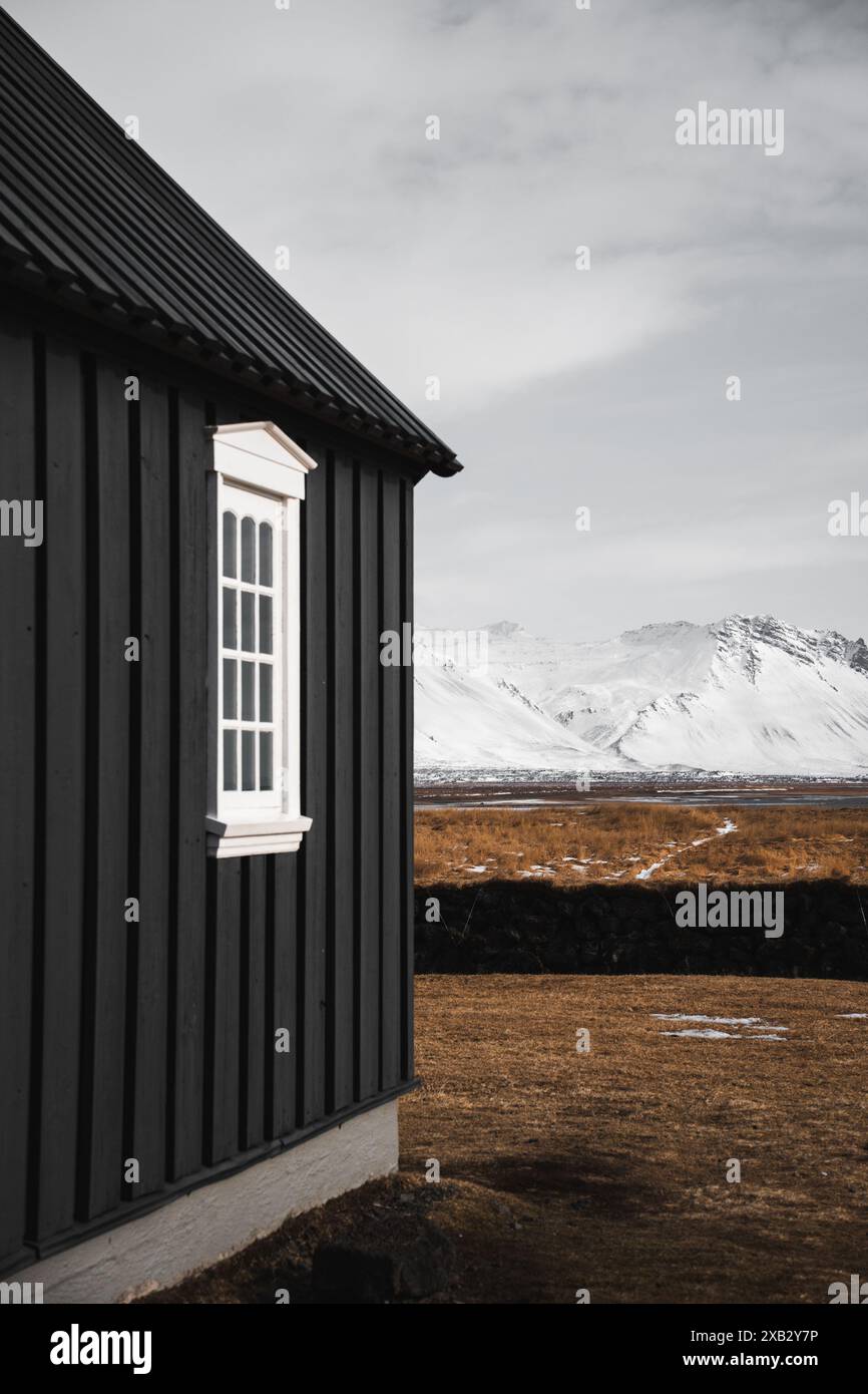 Ein malerisches, schwarzes traditionelles isländisches Cottage mit weißen Zierfenstern steht in starkem Kontrast zur Kulisse der schneebedeckten Berge auf dem Stockfoto