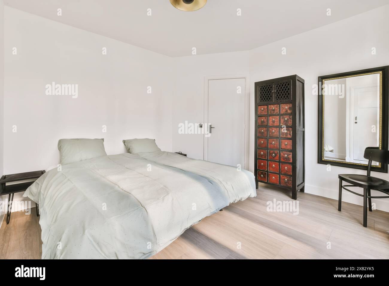Ein ruhiges, minimalistisches Schlafzimmer mit Twin-Betten mit hellem Bettzeug, schwarzem antiken Schrank mit roten Schubladen, großem Wandspiegel und Holzboden Stockfoto