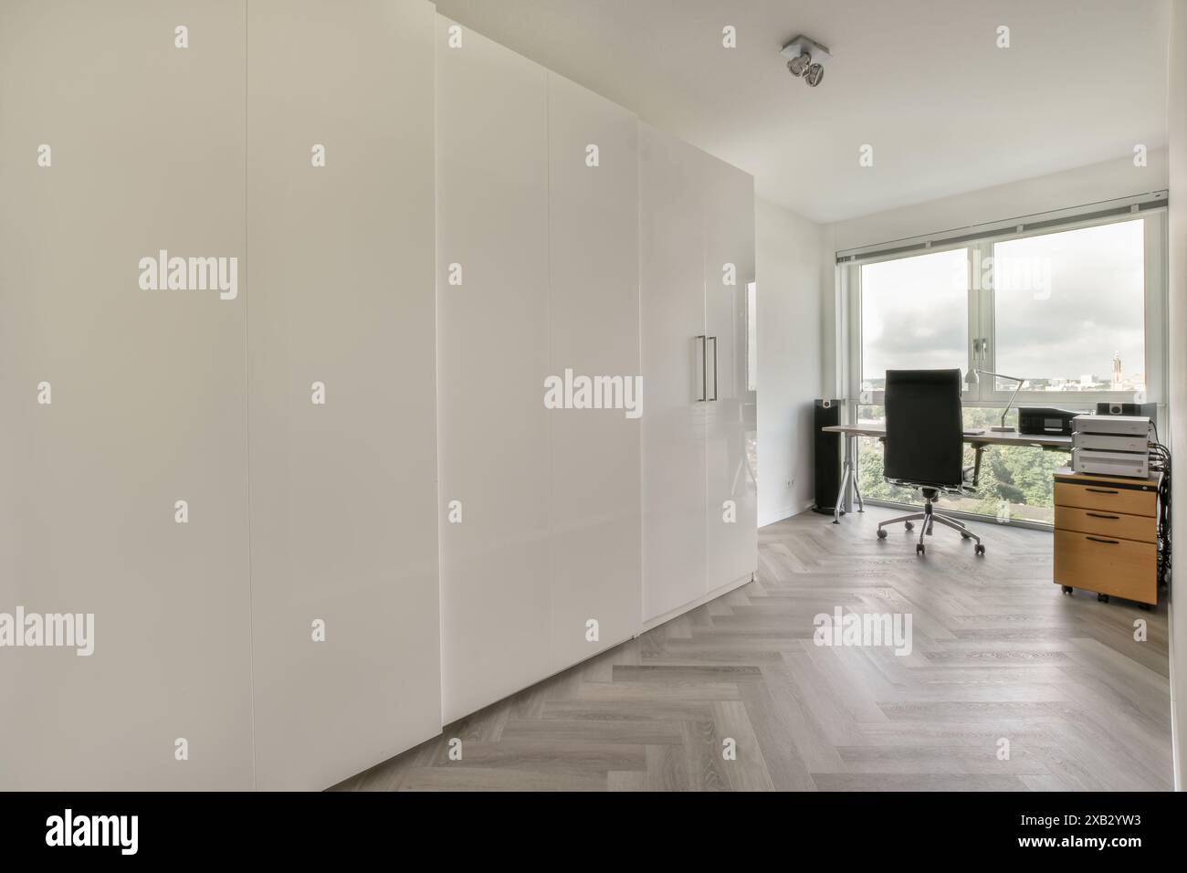 Ein helles und ordentliches, modernes Home Office-Interieur mit weißen Schränken, Fischgrätfußboden und großem Fenster mit malerischem Blick. Stockfoto