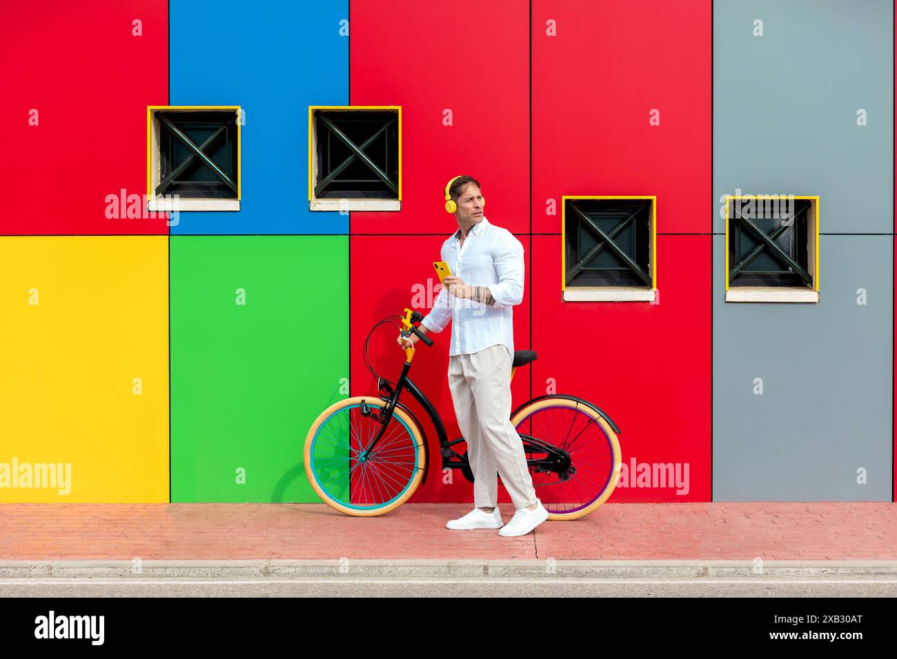 Ein Mann in lässiger Kleidung steht mit einem bunten Fahrrad vor einer lebhaft bemalten Wand mit quadratischen Fenstern Stockfoto