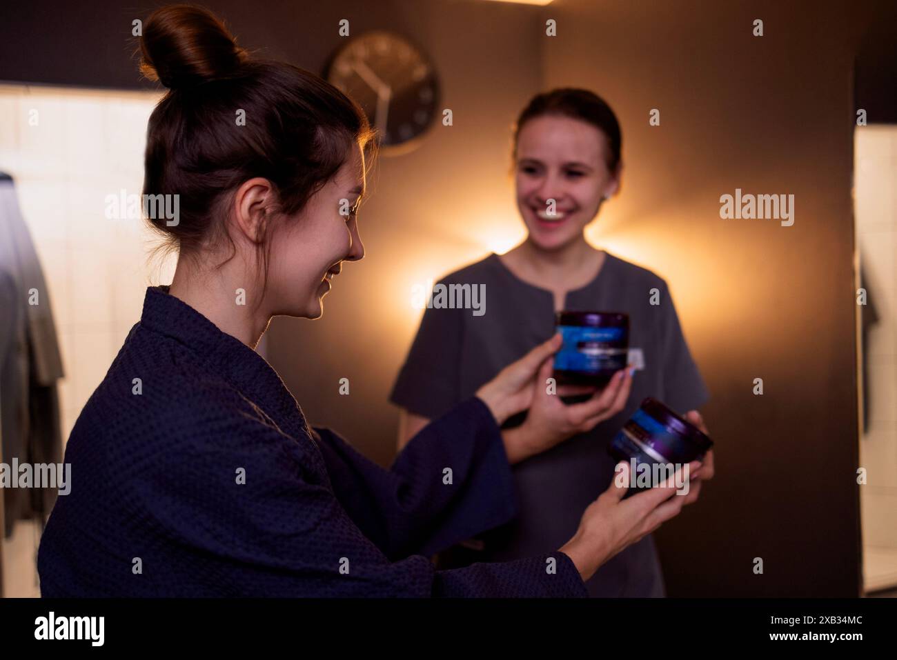 Eine junge Frau im Bademantel lächelt und wählt eine Creme im Spa. Die Kosmetikerin bietet ihrem attraktiven Kunden verschiedene Gesichtspflegeprodukte an Stockfoto