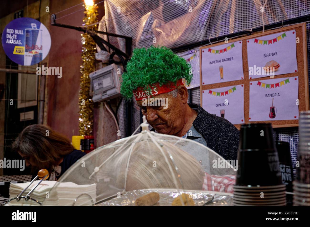 Ein reifer Mann mit grüner Perücke, der sich während der beliebten Heiligenfestlichkeiten in alfama intensiv in einem Essensstand konzentriert, ein farbenfroher illustriertes Menü hinter ihm Stockfoto