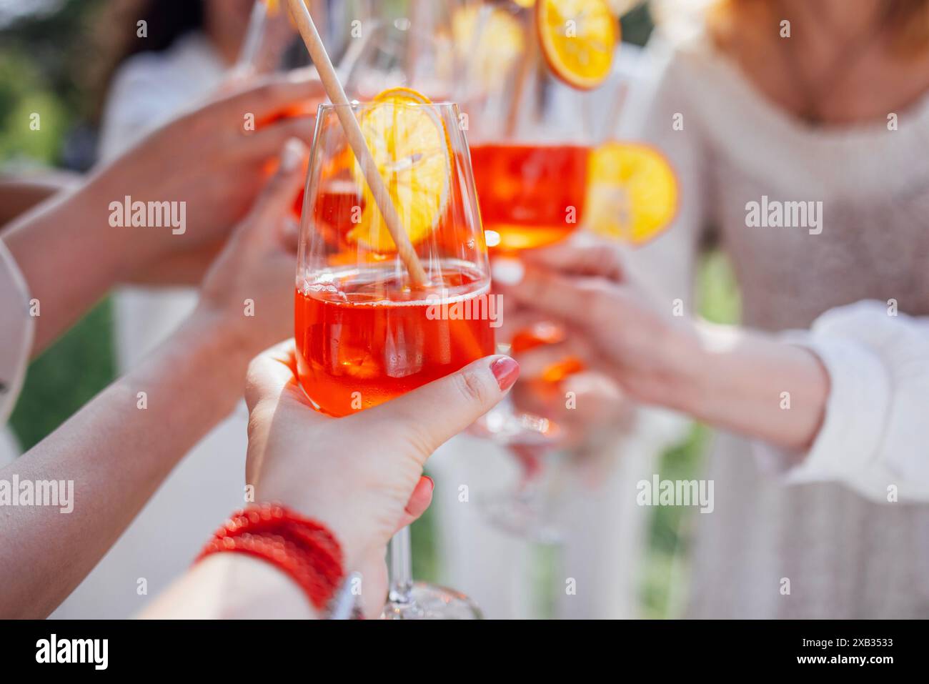 Nahaufnahme einer weiblichen Hand mit einer Brille mit einem köstlichen aperol. Junge Frauen in weißen Kleidern toasten mit orangen alkoholischen Getränken. Elegante Mädchen c Stockfoto