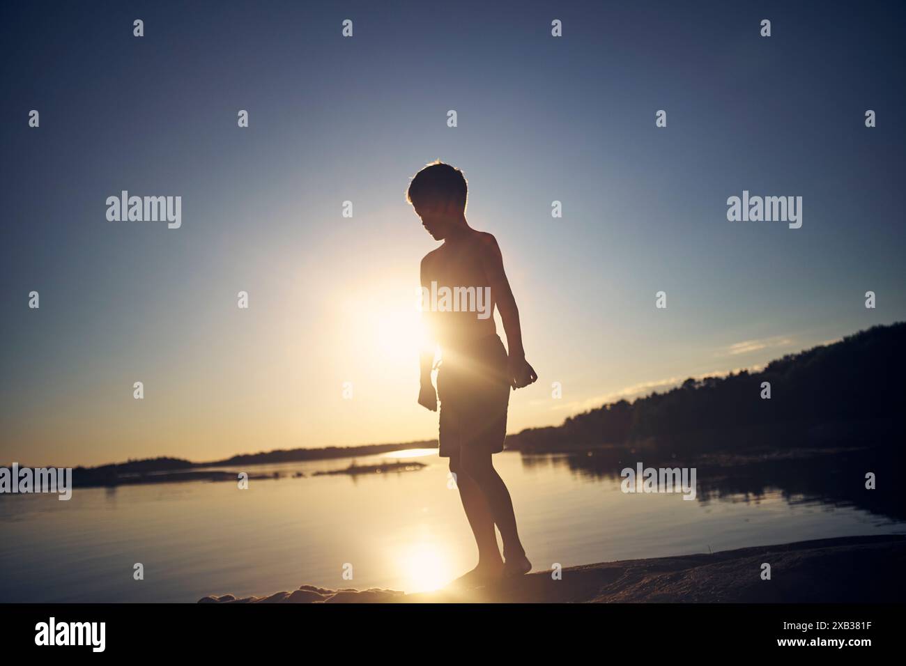 Die ganze Länge des Silhouettenjungen, der am Seeufer vor dem Himmel bei Sonnenuntergang steht Stockfoto