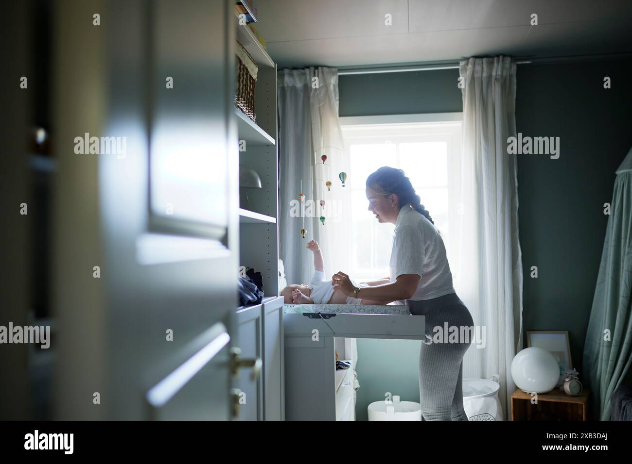 Seitenansicht der jungen Mutter, die die Windel des Babys wechselt, während sie am Fenster steht Stockfoto