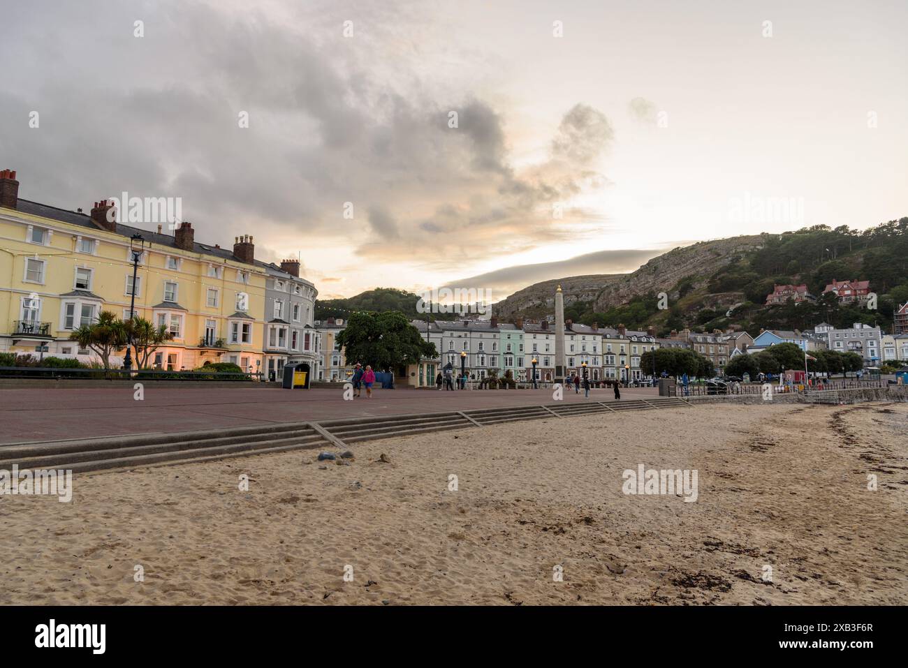 Menschen, die an der von pastellfarbenen Gebäuden gesäumten Promenade in der Küstenstadt Llandudno bei Sonnenuntergang spazieren Stockfoto