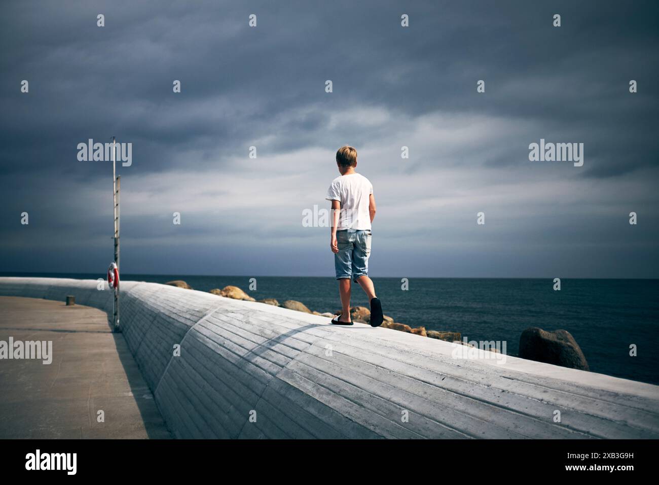 Rückansicht des Jungen, der auf einer Stützmauer in der Nähe des Meeres unter bewölktem Himmel läuft Stockfoto