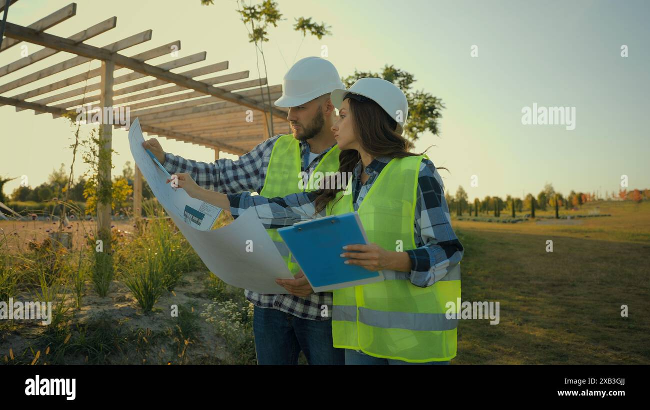 Kaukasische Bauunternehmer Ingenieure Kollegen weibliche Ingenieurinspektorin schreiben Bericht Hardhat männlicher Arbeiter Investor zeigen Bauplan Stockfoto